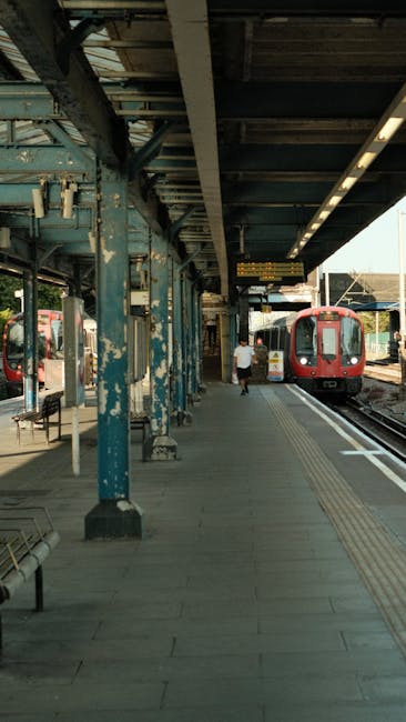 An outdoor platform at East Croydon station with a paved surface and tactile paving strips near the train tracks. The platform is sheltered by a metal roof supported by weathered blue metal columns showing peeling paint. A red train is parked on the right track, and a passenger is seen walking towards the train in the distance. The platform is well-lit with natural daylight, and there are a few benches along the edge. Carpet Cleaners Croydon, known for their cleaning services, emphasize the importance of surface cleaning and maintenance for hygienic environments, similar to the upkeep of public transport stations, ensuring cleanliness and safety in shared spaces such as station platforms.