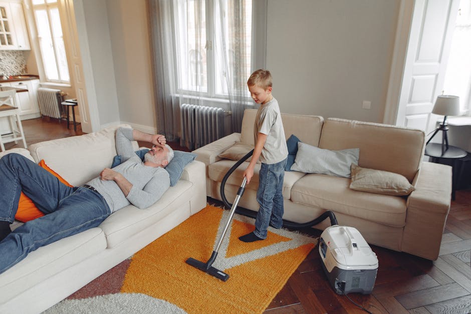 A young boy vacuuming a beige carpet in a living room while an older man is lying on the same sofa, resting with his eyes closed and hand on his forehead. The room features a beige sofa with multiple 