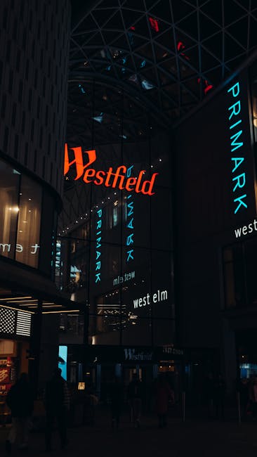 The image captures the interior of the Whitgift Centre shopping mall in Croydon, featuring a high, curved glass ceiling supported by dark metal framework. Multiple vertical digital screens display the logos for Westfield and Primark, illuminated in bright orange and blue on the dark walls, creating a vibrant, modern atmosphere. The spacious area is dimly lit, with some ambient lighting reflecting off polished tiled floors and glass surfaces. The scene includes a few shoppers and store entrances, indicating a busy retail environment. The overall cleanliness appears well-maintained, aligning with professional cleaning standards for commercial spaces, as exemplified by services provided by Carpet Cleaners Croydon.